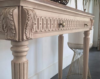 May include: A light beige console table with carved details and a dark brown top. The table features two drawers with brass knobs and is supported by fluted legs. Decorative orbs sit on top of the table.