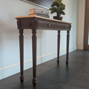 May include: A brown console table with a light-colored top, ornate carvings, and four fluted legs. The table has a drawer and is decorated with books and a small bonsai tree. The table is approximately 48 inches long.