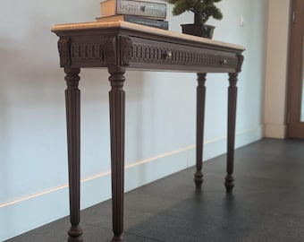 May include: A brown console table with a light-coloured top, ornate carvings, and four fluted legs. The table has a drawer and is decorated with books and a small bonsai tree. The table is approximately 122 cm long.