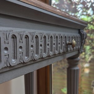 May include: A grey console table with a decorative carved drawer front and brass hardware. The tabletop has a dark wood finish. The table has four fluted legs. The table is approximately 76 cm long.