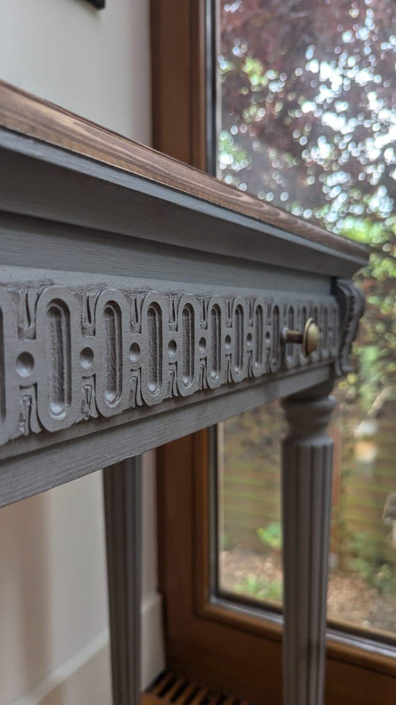 May include: A grey console table with a decorative carved drawer front and brass hardware. The tabletop has a dark wood finish. The table has four fluted legs. The table is approximately 76 cm long.