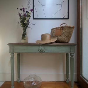 May include: A decorative console table with a distressed green finish, topped with a straw hat, woven basket, and a framed art print. A glass vase with flowers and a clear glass vase are also present. The table is 122 cm long.