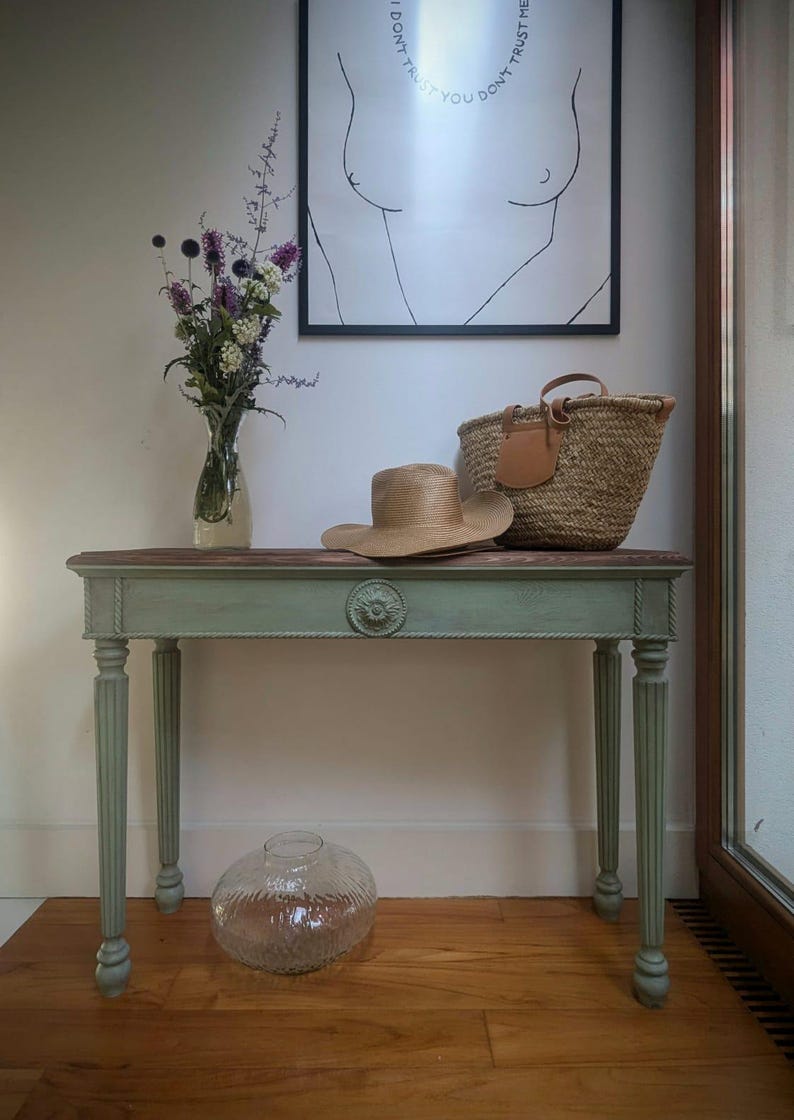 May include: A decorative console table with a distressed green finish, topped with a straw hat, woven basket, and a framed art print. A glass vase with flowers and a clear glass vase are also present. The table is 122 cm long.