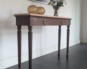 May include: A brown wooden console table with a light-coloured top, two drawers, and ornate legs. The table has gold-coloured drawer pulls and is decorated with two decorative glass spheres and a vase of flowers.