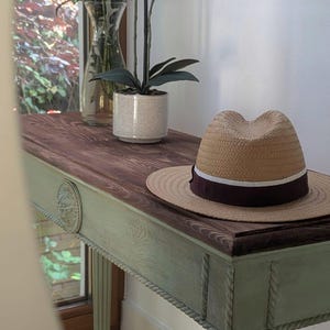 May include: A wooden console table with a brown top and a pale green base. A straw hat with a dark burgundy band rests on the table. A potted orchid and a vase of flowers are also present.