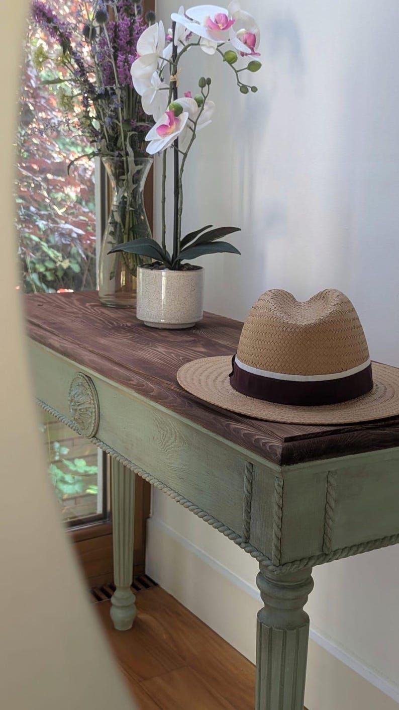 May include: A wooden console table with a brown top and a pale green base. A straw hat with a dark burgundy band rests on the table. A potted orchid and a vase of flowers are also present.