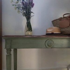 May include: A sage green console table with a dark wood top, adorned with a straw tote bag, a straw hat, and a vase of flowers. The table features carved details and fluted legs, adding a touch of elegance.