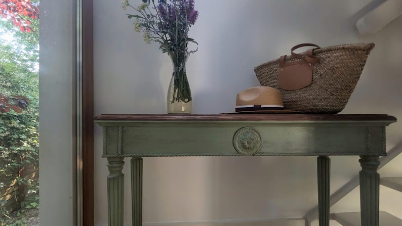 May include: A sage green console table with a dark wood top, adorned with a straw tote bag, a straw hat, and a vase of flowers. The table features carved details and fluted legs, adding a touch of elegance.