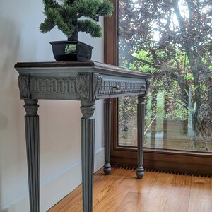 May include: A grey console table in an antique style, featuring a dark wood top and carved embellishments. A small bonsai tree in a black pot is placed on the table. The table has four fluted legs and a drawer with a brass knob. The table is in front of a window.