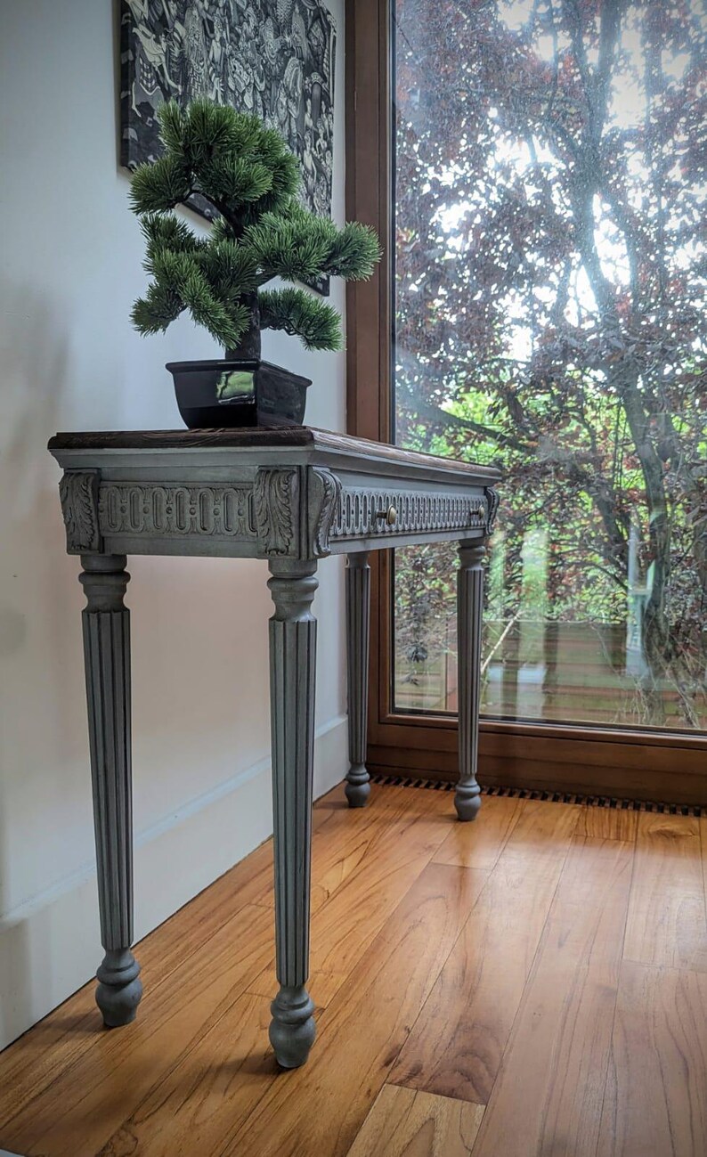 May include: A grey console table in an antique style, featuring a dark wood top and carved embellishments. A small bonsai tree in a black pot is placed on the table. The table has four fluted legs and a drawer with a brass knob. The table is in front of a window.