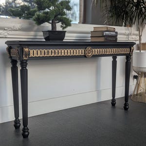 May include: A black console table with gold accents. The table has four slender, black legs and a rectangular top. A small bonsai tree in a black pot sits on the table, along with a stack of books. The table's trim features a gold chain-link design.