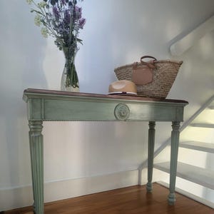 May include: A sage green console table with a brown top, featuring a straw hat, woven tote bag, and a vase of purple and white flowers. The table has decorative carved details and fluted legs.