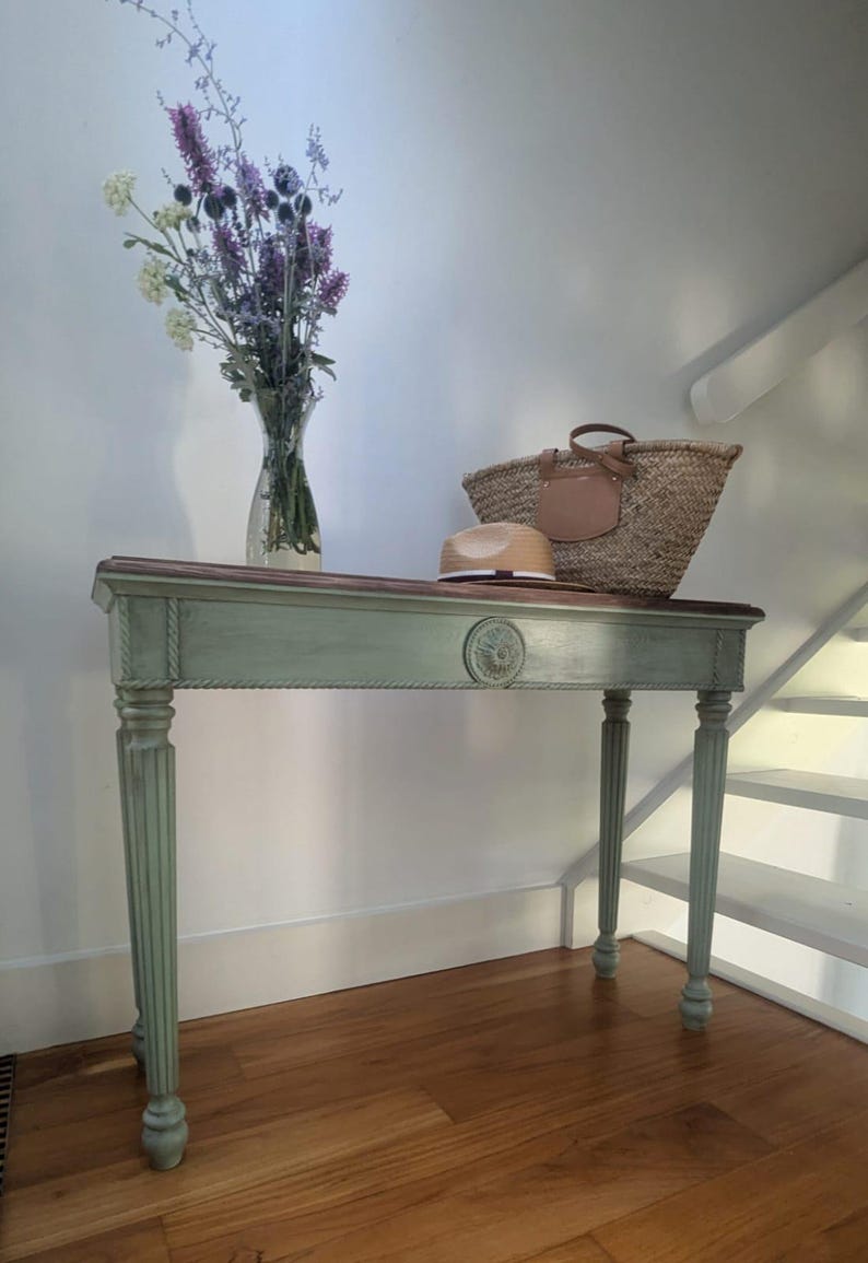 May include: A sage green console table with a brown top, featuring a straw hat, woven tote bag, and a vase of purple and white flowers. The table has decorative carved details and fluted legs.
