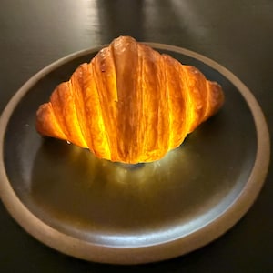 Puede incluir: Un cruas&aacute;n dorado sobre un plato verde oscuro con un borde marr&oacute;n claro. La pasta hojaldrada est&aacute; iluminada, resaltando sus capas. El plato descansa sobre una superficie oscura.