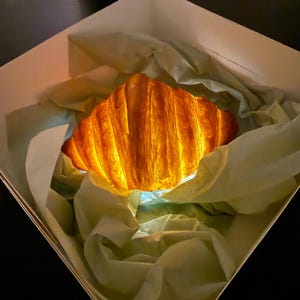 Puede incluir: Un cruas&aacute;n dorado y crujiente, iluminado desde dentro, descansa en una caja blanca forrada con papel de seda verde claro. Las capas de la pasteler&iacute;a son visibles, creando una apariencia texturizada. La imagen est&aacute; bien iluminada, resaltando los detalles del cruas&aacute;n.
