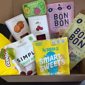 Pode incluir: Uma caixa de cartão cheia de vários produtos de doces e gomas. Os pacotes incluem 'Smart Sweets' com 3g de açúcar, 'Simply Sours', gomas 'Bon Bon' e 'Jocelyn & Co' fruit sours. A embalagem apresenta cores vivas e ilustrações de frutas.