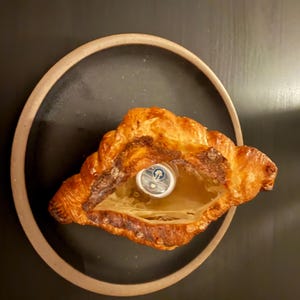 Puede incluir: Un cruas&aacute;n dorado y crujiente con un centro ahuecado, que revela un peque&ntilde;o objeto plateado. La pasteler&iacute;a est&aacute; colocada sobre un plato oscuro con un borde marr&oacute;n claro, sobre un fondo oscuro. La imagen destaca la textura y el color del producto horneado.