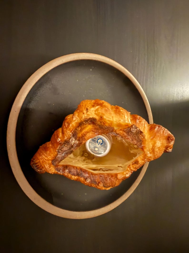 Puede incluir: Un cruas&aacute;n dorado y crujiente con un centro ahuecado, que revela un peque&ntilde;o objeto plateado. La pasteler&iacute;a est&aacute; colocada sobre un plato oscuro con un borde marr&oacute;n claro, sobre un fondo oscuro. La imagen destaca la textura y el color del producto horneado.