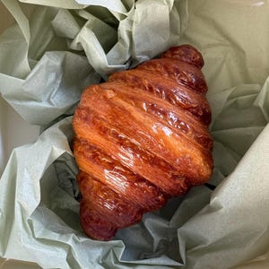 Puede incluir: Un cruas&aacute;n dorado y brillante, con una superficie caramelizada, se presenta en una caja blanca forrada con papel de seda verde p&aacute;lido. Las capas de la pasteler&iacute;a son visibles, sugiriendo una textura hojaldrada. Vista superior, destacando la forma y el color.