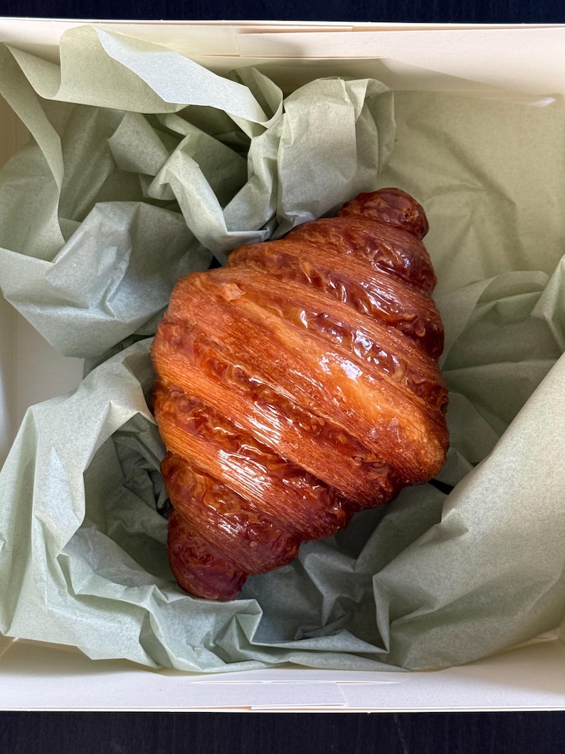 Puede incluir: Un cruas&aacute;n dorado y brillante, con una superficie caramelizada, se presenta en una caja blanca forrada con papel de seda verde p&aacute;lido. Las capas de la pasteler&iacute;a son visibles, sugiriendo una textura hojaldrada. Vista superior, destacando la forma y el color.
