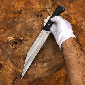 Könnte beinhalten: Ein großes Messer mit silberner Klinge und schwarzem Griff. Das Messer wird von einer behandschuhten Hand gehalten, wobei die Klinge auf den Betrachter zeigt. Auf der Klinge steht der Text "RANDOM BLADES". Das Messer liegt auf einer Holzoberfläche.