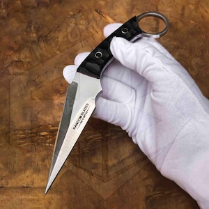 May include: A silver and black dagger with a ring on the handle. The blade is sharp and pointed, with the text "RANDOM BLADES" printed on the side. The handle is black with a textured grip.
