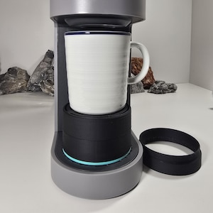 Op de afbeelding: Een grijze Keurig koffiemachine met een witte mok erin. De mok heeft een blauwe rand en horizontale lijnen. De merknaam Keurig staat op de voorkant. Een zwarte ring ligt naast het koffiezetapparaat.