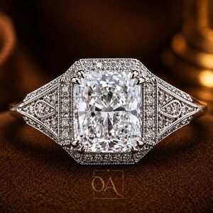 Puede incluir: Un anillo de diamantes con una piedra central rectangular de talla radiante, rodeada por un halo de diamantes m&aacute;s peque&ntilde;os. El anillo presenta intrincados detalles en la banda. La montura es de metal brillante, posiblemente platino u oro blanco, sobre un fondo marr&oacute;n.