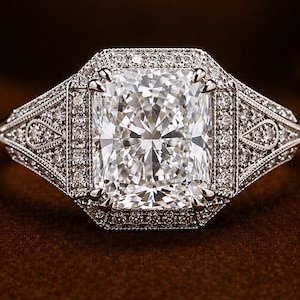 Puede incluir: Un anillo de diamantes de talla rectangular con un halo de diamantes m&aacute;s peque&ntilde;os. El anillo presenta un dise&ntilde;o intrincado en la banda y alrededor de la piedra central. El anillo est&aacute; sobre una superficie marr&oacute;n, con las letras "OA" en un cuadrado dorado en el fondo.