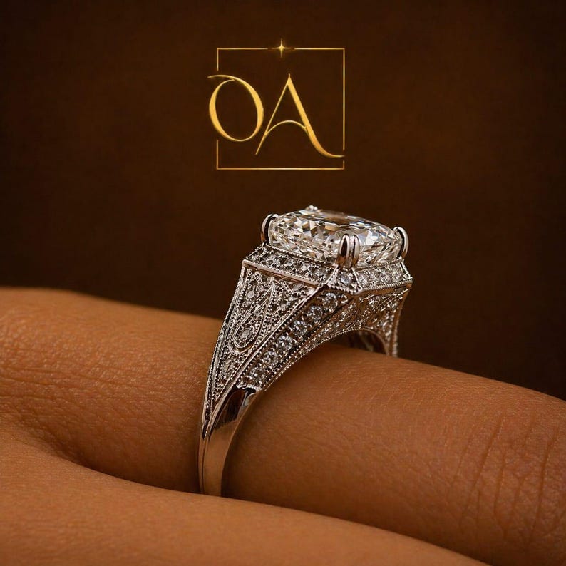 Puede incluir: Un anillo detallado, plateado, con una gran piedra central de diamante de talla cuadrada. El anillo presenta intrincados filigranas y peque&ntilde;as piedras de acento, sobre un fondo marr&oacute;n oscuro. El anillo est&aacute; en un dedo.