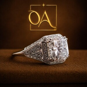 Puede incluir: Primer plano de un anillo de diamantes con una gran piedra central de talla cuadrada. El anillo est&aacute; engastado en una banda plateada con detalles intrincados y diamantes de acento m&aacute;s peque&ntilde;os. El fondo es marr&oacute;n.