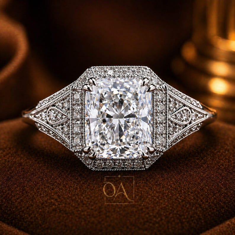 Puede incluir: Un anillo de diamantes con una piedra central rectangular de talla radiante, rodeada por un halo de diamantes m&aacute;s peque&ntilde;os. El anillo presenta intrincados detalles en la banda. La montura es de metal brillante, posiblemente platino u oro blanco, sobre un fondo marr&oacute;n.