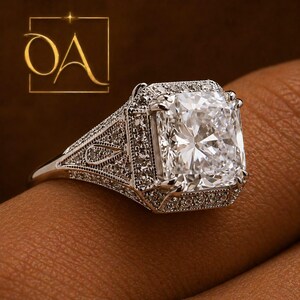 Puede incluir: Primer plano de un anillo de diamantes con una gran piedra central de talla cuadrada. El anillo tiene un engaste detallado y ornamentado con diamantes de acento m&aacute;s peque&ntilde;os. La banda es plateada, sobre un fondo marr&oacute;n.