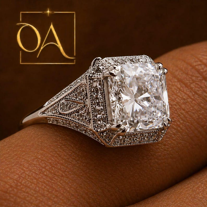 Puede incluir: Primer plano de un anillo de diamantes con una gran piedra central de talla cuadrada. El anillo tiene un engaste detallado y ornamentado con diamantes de acento m&aacute;s peque&ntilde;os. La banda es plateada, sobre un fondo marr&oacute;n.