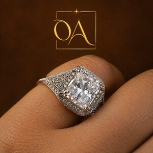 Puede incluir: Primer plano de un anillo de diamantes con una gran piedra central de talla cuadrada. El anillo tiene un engaste detallado de estilo vintage con diamantes m&aacute;s peque&ntilde;os. El anillo est&aacute; en un dedo, con un fondo marr&oacute;n y las letras "OA" en un cuadrado dorado.