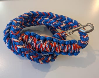 May include: Braided dog leash in blue, red, and white. The leash is made of thick rope and features a silver-colored metal clasp. The leash is coiled on a light-colored surface.