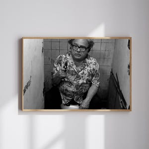 May include: Black and white framed print featuring a man in glasses and a floral shirt holding a gun. The man is seated in a bathroom stall. The print is in a wooden frame.