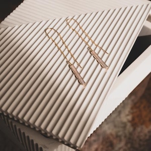 May include: A pair of gold-toned threader earrings featuring a delicate, wavy chain design that connects to a slender, rectangular bar pendant at the base. The earrings have a polished metallic finish and a long, dangling silhouette.