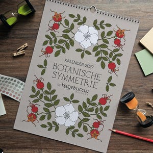 Könnte beinhalten: Ein Wandkalender für 2027 mit dem Titel Botanische Symmetrie by Hagebuttin Designs, der ein hellgraues Deckblatt mit einer botanischen Illustration aus weißen Blüten, grünen Blättern und roten Hagebutten in symmetrischer Anordnung zeigt.