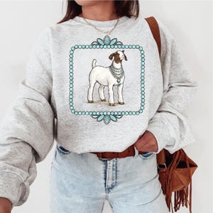 May include: Light gray sweatshirt featuring a goat illustration within a turquoise frame. The goat is white with brown markings and wears a pearl necklace. The frame has a beaded border and floral accents. The sweatshirt has long sleeves.