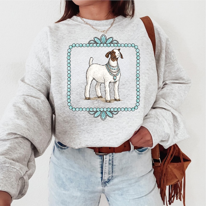 May include: Light gray sweatshirt featuring a goat illustration within a turquoise frame. The goat is white with brown markings and wears a pearl necklace. The frame has a beaded border and floral accents. The sweatshirt has long sleeves.