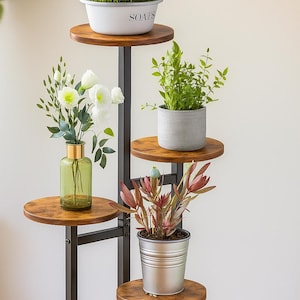 May include: A multi-tiered plant stand with a black metal frame and wooden shelves. The stand displays various potted plants, including herbs in a white container labeled "SOAPS", white flowers in a green vase, and succulents.