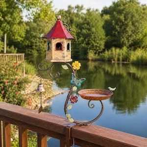 May include: A decorative bird feeder and water dish set. The birdhouse has a red roof and is supported by a metal bracket with floral and butterfly accents. A small bell hangs below the birdhouse. The water dish is filled with water.