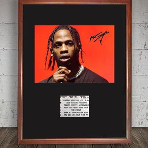 May include: Framed print featuring a portrait of Travis Scott against a red backdrop, with his signature. Below the image is a concert ticket with the text "TRAVIS SCOTT: ASTROWORLD WISH YOU WERE HERE TOUR". The frame is brown.