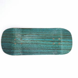 May include: A teal and black fingerboard deck with a wood grain pattern. The miniature skateboard has a rounded shape with pre-drilled holes for mounting trucks. The deck is a vibrant teal color with dark black lines running across it.