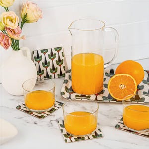 Puede incluir: Una jarra y vasos de vidrio transparente llenos de zumo de naranja, junto con naranjas frescas. La escena incluye posavasos decorativos y una bandeja con un estampado floral verde y marrón, y un jarrón blanco con flores.