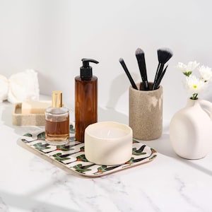 Könnte beinhalten: Ein rechteckiges Tablett mit einem Blattmuster hält eine Parfümflasche, einen braunen Seifenspender, eine Kerze und eine kleine Vase mit weißen Blumen. Make-up-Pinsel befinden sich in einem beigefarbenen Halter.