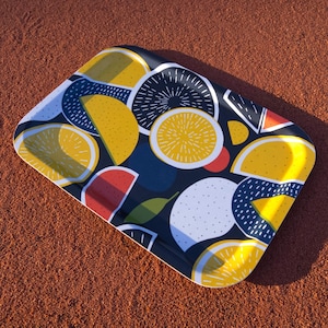Pode incluir: Bandeja retangular com um padrão colorido de frutas cítricas. O design apresenta limões amarelos, gomos de laranja e círculos brancos pontilhados em um fundo azul escuro. A bandeja tem um acabamento brilhante.
