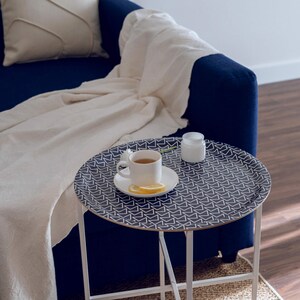 Könnte beinhalten: Ein runder Beistelltisch mit einem gemusterten Tablett, das eine Tasse Tee, eine Untertasse und eine Zitronenscheibe enthält. Das Tablett hat einen marineblauen Hintergrund mit einem weißen geometrischen Design. Neben der Tasse steht ein weißes Glas. Der Tisch hat weiße Beine.