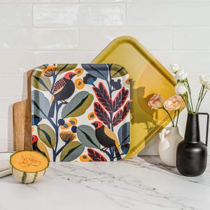 Könnte beinhalten: Ein quadratisches Serviertablett mit Vogel- und Blumendesign, ein gelbes Tablett und ein Holzschneidebrett werden auf einer weißen Oberfläche präsentiert. Eine schwarze Vase, eine weiße Vase und eine halbe Cantaloupe-Melone sind ebenfalls vorhanden.